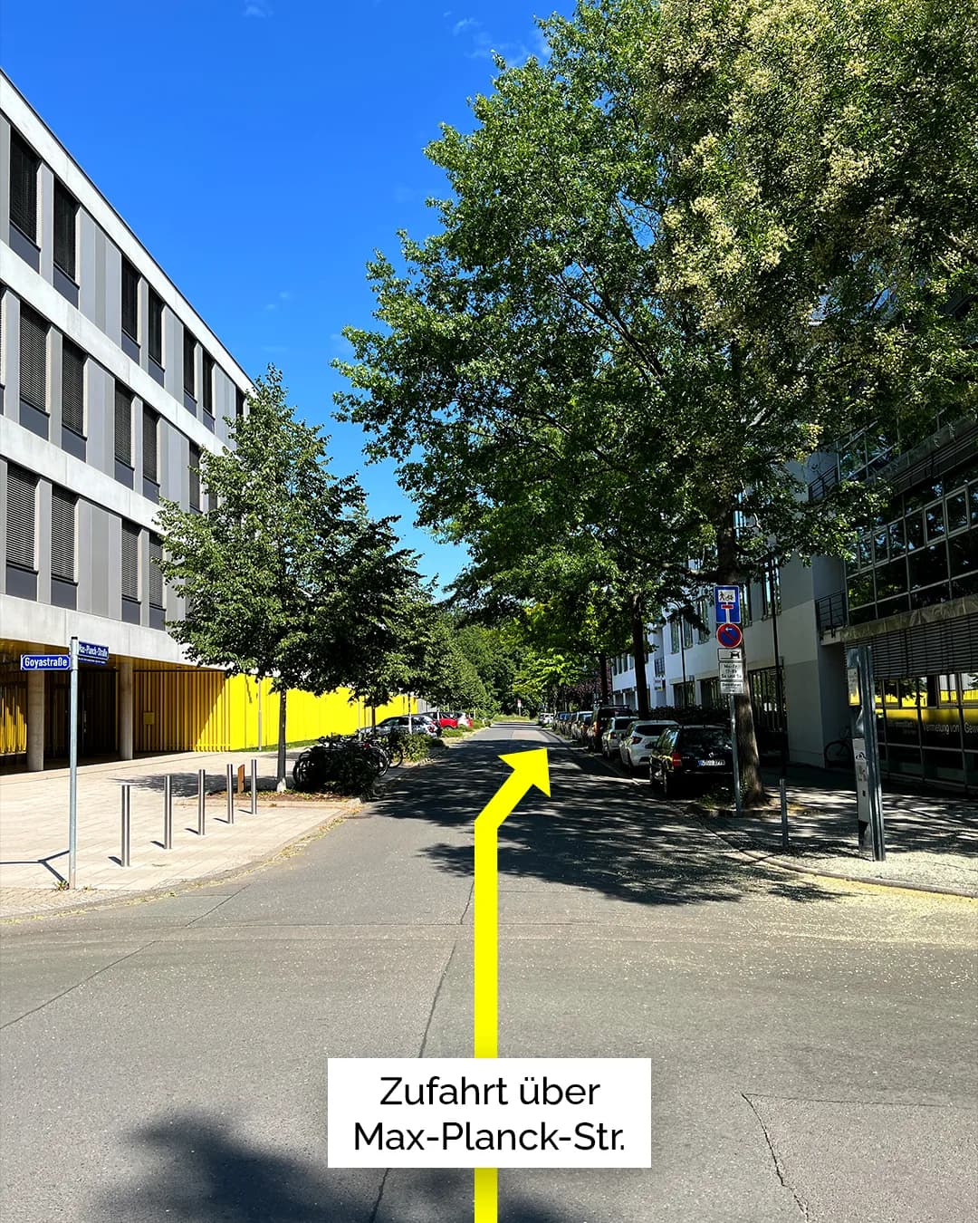 Straßenansicht mit Blick entlang einer ruhigen, von Bäumen gesäumten Straße. Ein gelber Pfeil auf der Fahrbahn weist geradeaus, darunter steht der Hinweis „Zufahrt über Max-Planck-Str.“. Am Straßenrand sind Parkplätze und abgestellte Fahrzeuge zu sehen, links befindet sich ein modernes Gebäude mit gelber Fassade.