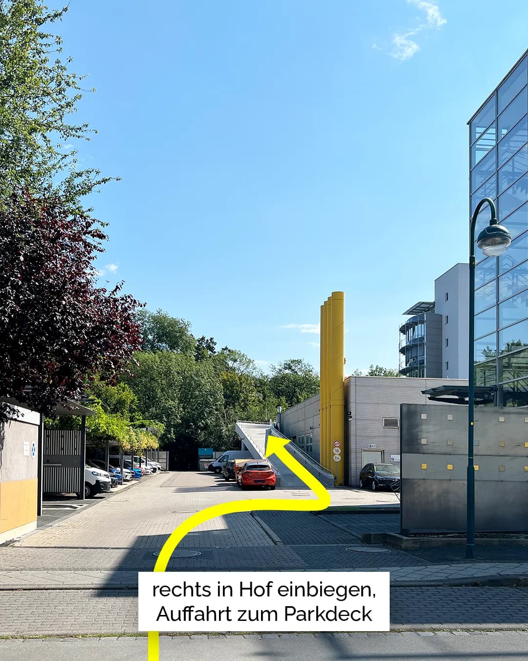 Zufahrt zu einem Innenhof mit Parkmöglichkeiten und Auffahrt zu einem Parkdeck. Ein gelber Pfeil markiert die Fahrtrichtung nach rechts in den Hof, begleitet vom Hinweis „rechts in Hof einbiegen, Auffahrt zum Parkdeck“. Mehrere Fahrzeuge sind entlang der Zufahrt und unter einem Carport geparkt.