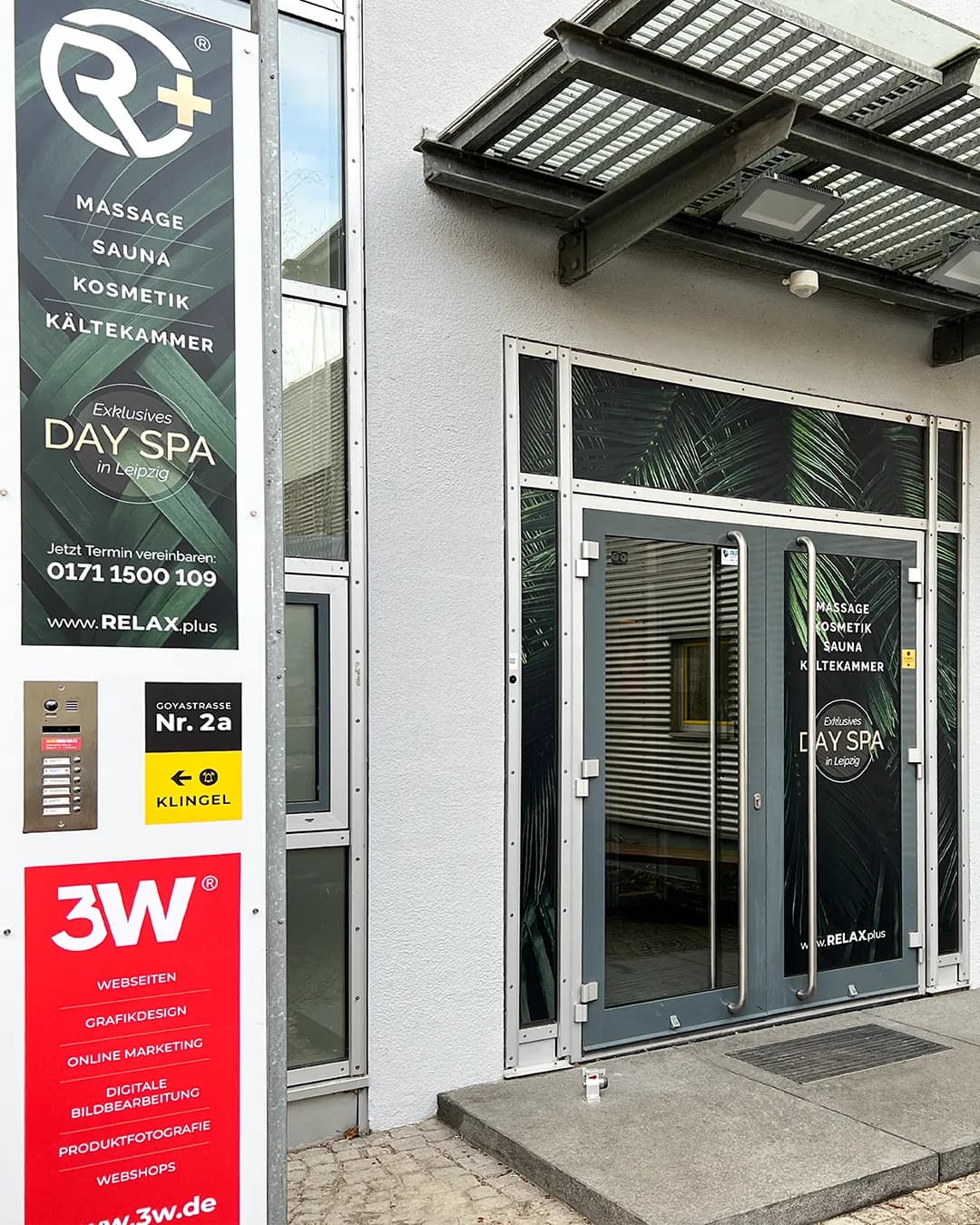 Eingangsbereich eines Day Spas in einem modernen Gebäude mit Glasdoppeltür und großem Werbebanner. Auf der linken Seite ist ein vertikales Schild mit der Aufschrift „Massage, Sauna, Kosmetik, Kältekammer – Exklusives Day Spa in Leipzig“ sowie Kontaktdaten zu sehen. Neben der Tür befinden sich weitere Firmenschilder, darunter „3W“.